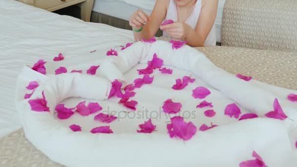 Una niña de 6 años con un brazalete de hotel en la mano está jugando con pétalos de flores. En su cama hay un corazón de toallas - un cumplido del hotel, excelente servicio — Vídeo de stock