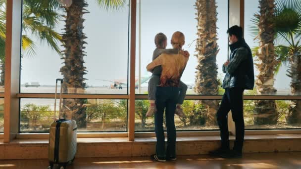 Una familia de tres personas mira a la gran ventana de la terminal del aeropuerto. Admira los aviones, el sol brilla fuera de la ventana y las palmeras crecen . — Vídeos de Stock