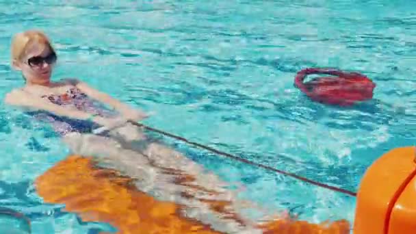 Una joven con gafas de sol está entrenando músculos en un simulador en la piscina. Actividades activas al aire libre — Vídeo de stock