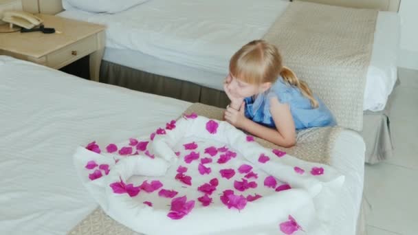 Una niña de 6 años con un brazalete de hotel en la mano está jugando con pétalos de flores. En su cama hay un corazón de toallas - un cumplido del hotel, excelente servicio — Vídeo de stock