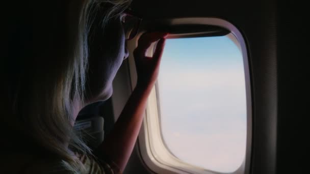 若い女性は、飛行機の窓と窓の外に見えるを閉じます。カーテンの開口を有するダーク フレームが点灯します。 — ストック動画