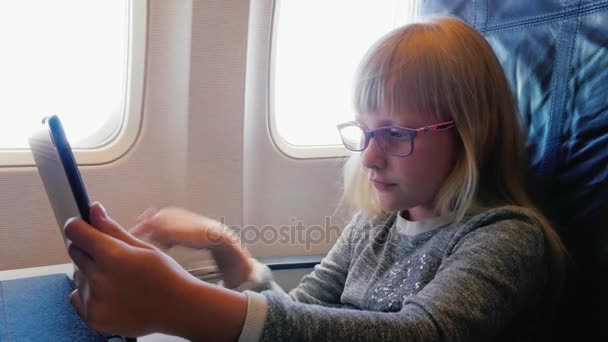 En tjej med glasögon i 6 år reser med flyg. Använder en digital tablett under flygning — Stockvideo
