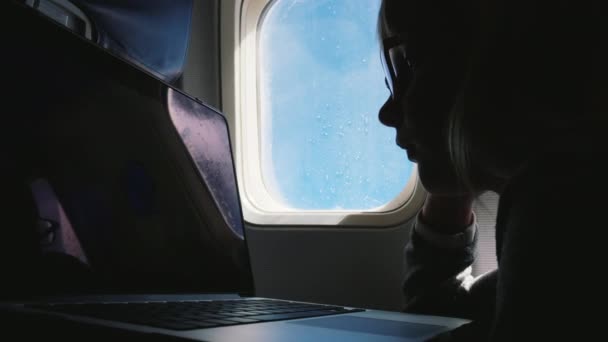 Silhouette eines Mädchens mit Brille, das auf den Laptop-Bildschirm blickt. fliegt im Flugzeug — Stockvideo
