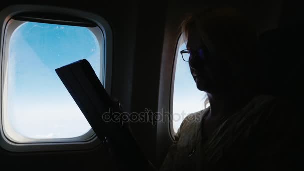 Eine Frau fliegt im Flugzeug und liest etwas auf dem Tablet. Silhouette, Seitenansicht — Stockvideo