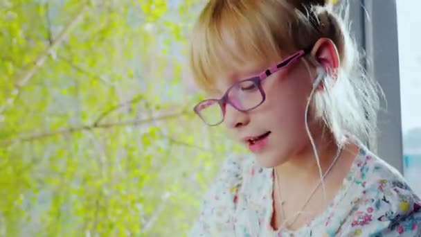 Chica divertida en los auriculares escuchando música, cantando. Se sienta en el alféizar de la ventana contra la ventana, fuera del soleado día de primavera. Excelente estado de ánimo — Vídeo de stock