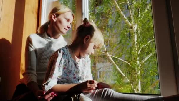 妈妈和女儿是 6 岁，坐在窗台上，看着窗外。窗外是春天，在树上的绿叶，阳光明媚。概念-等待某个人，梦想 — 图库视频影像