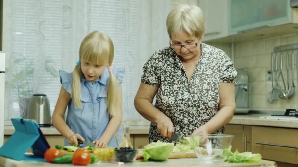 Samen koken. Het meisje van 6 jaar helpt haar oma in de keuken, kijken naar het recept van de salade op de Tablet PC — Stockvideo