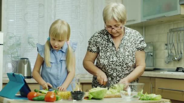 Společně vaříme. Dívka 6 let pomáhá babička v kuchyni, sledování salát recept na tabletu — Stock video