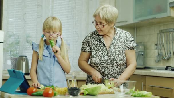 Chica divertida 6 años de edad ayuda a su abuela a preparar comidas en la cocina. Come un pepino, diviértanse juntos. Alimentación saludable — Vídeos de Stock