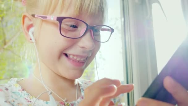 La chica graciosa sonríe y usa el teléfono. Lleva gafas de color rosa y auriculares blancos. Retrato de un niño feliz con un dispositivo — Vídeo de stock