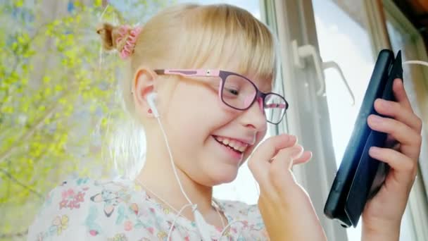 Retrato alegre chica rubia en gafas de color rosa con un teléfono móvil. Se ríe, juega por teléfono. Detrás de su ventana y el sol primaveral brilla — Vídeos de Stock