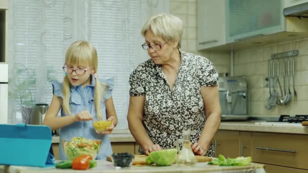 Шестилетняя девочка в розовых очках готовит салат на кухне. Ее бабушка помогает, использовать планшет — стоковое видео
