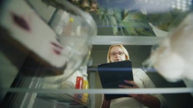 Online sipariş gıda. Genç bir kadın için bir bakkal alışveriş listesini yapar. Buzdolabında yiyecek bakar, bir tablet kullanır. İç görünüm