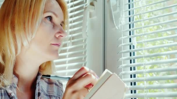 Молодая женщина пишет в блокноте. Он сидит у окна с открытыми жалюзи. Мечты, планы на будущее — стоковое видео