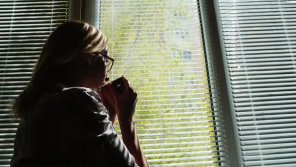 Silueta de una mujer joven junto a la ventana. Mira la calle a través de las persianas, bebe café de una taza. Mira hacia adelante — Vídeos de Stock