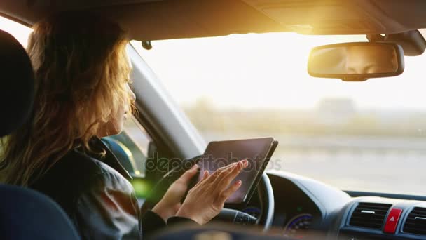 Una giovane donna gode di un tablet in macchina, navigando e orientandosi in un luogo sconosciuto. Vista posteriore al tramonto — Video Stock