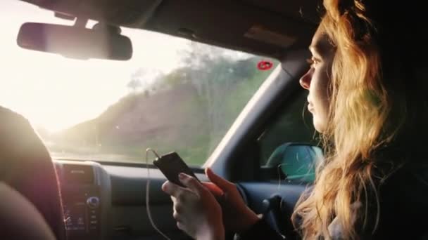 Nos gusta viajar. Una mujer joven con un teléfono monta en el asiento del pasajero delantero del coche. En los rayos de un sol poniente — Vídeos de Stock