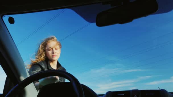 Een aantrekkelijke vrouw wrijft het glas van haar auto op het platteland. Tegen de achtergrond van de blauwe hemel — Stockvideo