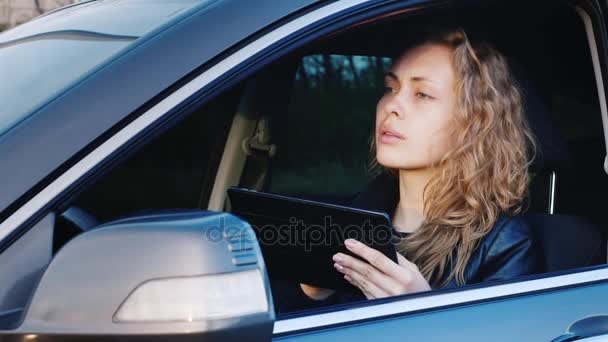 Mujer ligeramente alarmada utiliza una tableta, se sienta al volante del coche. Encuentra el camino correcto, navega en tránsito. Vista frontal — Vídeo de stock