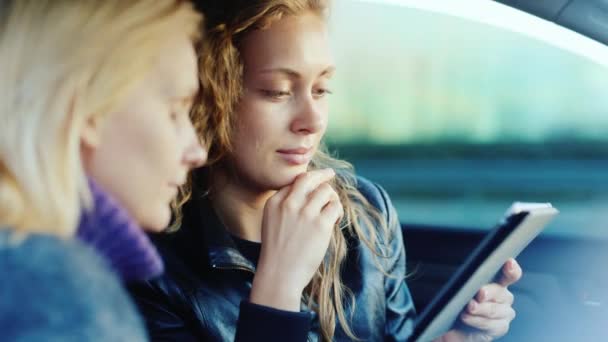 Dos mujeres se comunican dentro del coche. Ríete, sonríe, disfruta de la tableta. Vista lateral — Vídeo de stock