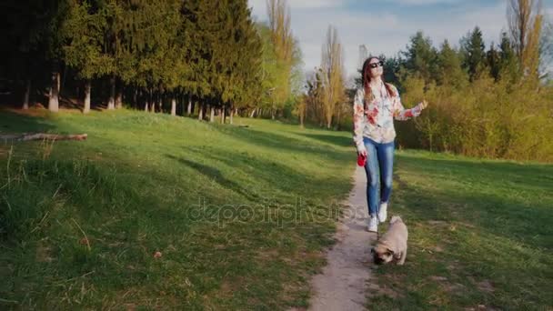 Молодая женщина в солнечных очках гуляет с собакой в парке. Идет на фоне живописной природы вдоль тропы — стоковое видео