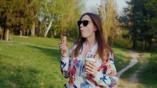 A young woman walks in the park, eats a food and drinks coffee from a paper cup. Coffee and hotdog - snack on the go — Stock Video