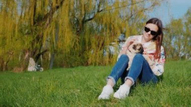 Yeşil çimenlerin üzerinde oturan parkta oturan genç kadın. Kucağında pug köpek yavrusu tutar, bir köpekle oynar. Eğlenir