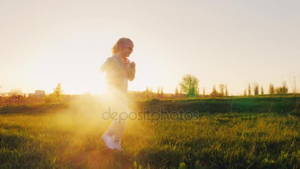 ピンクのメガネで面白い女の子とスポーツは体操に合います。夕暮れ時を緑の牧草地を通って実行している、太陽が後ろから美しく輝いています。 — ストック動画