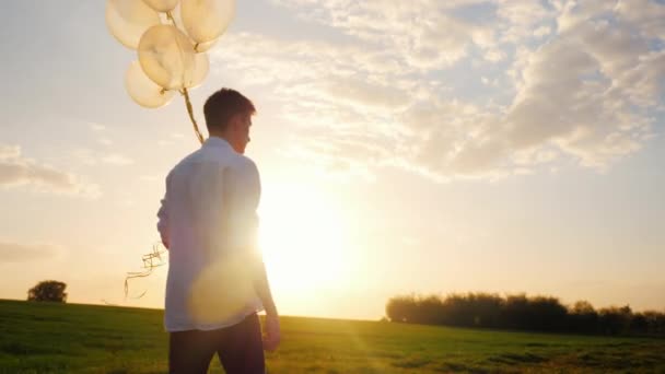 Tonåring i en vit smart skjorta med ballonger i handen. Går i en pittoresk plats mot solnedgången. Steadicam skott — Stockvideo