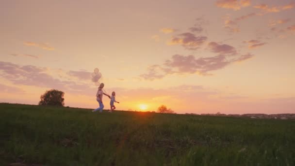 Mamá y su hija están corriendo por un prado verde con globos. Juntos pasar un buen rato — Vídeos de Stock