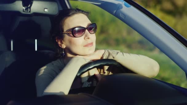 Una mujer en gafas de sol se relaja en el coche. Se sienta detrás del volante, admirando el hermoso paisaje al atardecer. Conductor feliz del concepto — Vídeos de Stock