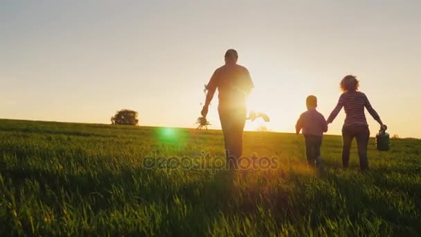 Visão traseira: Uma família amigável com um filho jovem vai plantar uma árvore. Leve uma planta cultivada de sementes, pá e regador. Silhuetas em um belo campo em um fundo de pôr do sol — Vídeo de Stock