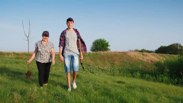 Una anciana con un nieto adolescente va a plantar un árbol. Llevan en sus manos herramientas de jardín y una plántula de árbol. Comunicación de generaciones, personas mayores activas — Vídeos de Stock