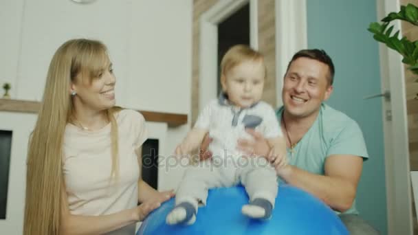 Jovens pais caucasianos brincam com seu bebê. Rolá-lo em uma bola de massagem — Vídeo de Stock