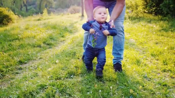 Grappig jongetje leert te lopen. Zijn handen worden ondersteund door zijn vader. De jongen lacht, een Lila bloem in zijn handen houdt. Slow motion video — Stockvideo