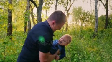 Mutlu bir genç baba bebek oğlu ile oynuyor. Yeşil ağaçlar yakınındaki güzel bir yerde, güzel güneş parlıyor