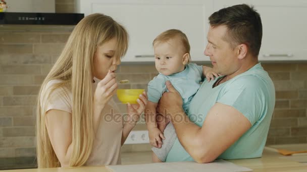 Papá y mamá alimentan a su pequeño hijo juntos. Un hombre sostiene a un niño sobre sus gallinas, su madre lo alimenta con una cuchara. Comida para bebés, familia feliz — Vídeos de Stock