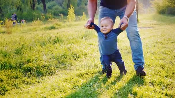 Um rapaz de um ano com um sorriso nos lábios. Aprende a andar no parque. Ele é apoiado por Papas mãos. Vídeo em câmera lenta — Vídeo de Stock
