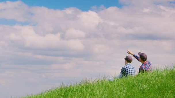 Два подростка в шапочках сидят на зеленом холме на фоне голубого неба. Они говорят и поднимают руки к небу. Детские мечты, идиллические пейзажи. Копикосмическая композиция — стоковое видео