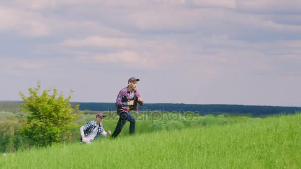 Zwei Teenager knallen nacheinander über einen schönen grünen Hügel vor blauem Himmel mit Wolken — Stockvideo