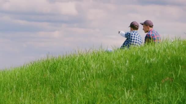 Dva mladí kluci sedí na vrcholu zeleného kopce, komunikovat a snít. Na pozadí krásná modrá obloha s mraky. Copyspace složení — Stock video