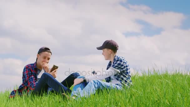 Dos adolescentes se comunican en la naturaleza. Se sientan en un prado verde, usan teléfonos. En el fondo de un hermoso cielo — Vídeos de Stock