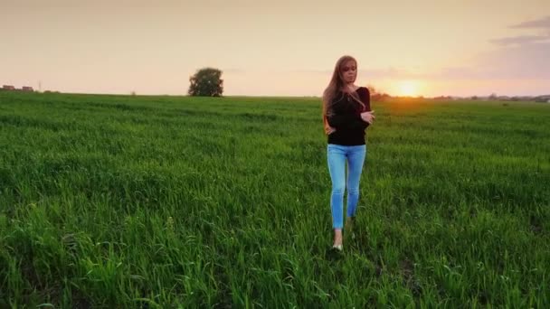 Fiatal kaukázusi nő séta egy zöld rét a naplemente. Átgondolt, magába foglalja magát a kezét. Lassú mozgás Steadicamnél videóinak — Stock videók