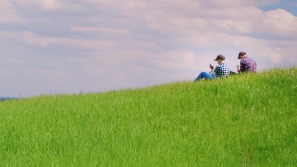 Dois adolescentes usam telefones celulares em um lugar pitoresco em uma colina verde. Uma paisagem serena - tecnologia e estar em contato em qualquer lugar. Composição do espaço de cópia — Vídeo de Stock