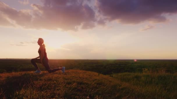 Une femme fait du sport dans un endroit pittoresque au coucher du soleil. Sur fond de ciel et de plaines verdoyantes, au sommet de la colline. Steadicam shot — Video