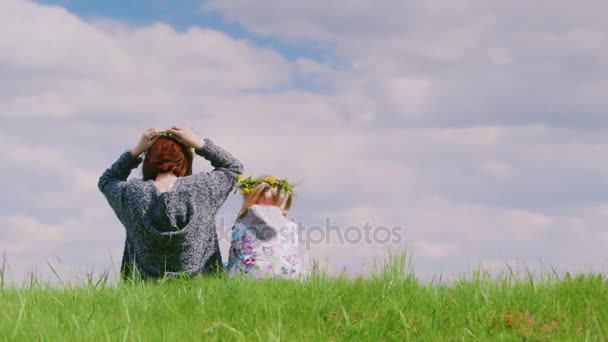 Вид сзади: женщина средних лет со своей дочерью венчает вместе венок из диких цветов. На зеленом холме на фоне голубого неба — стоковое видео
