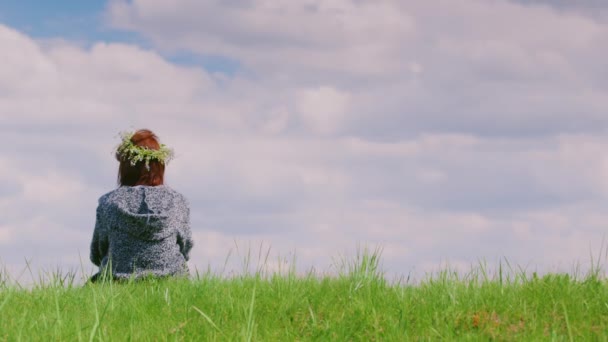 孤独な女性は、緑の丘の上に座っています。彼女の頭の上には、花の花輪です。Copyspace 組成の背面ビュー — ストック動画