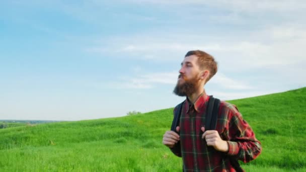 Um jovem hipster barbudo com uma mochila atrás dele. Vai ao longo do vale verde contra o céu azul. Adoro viajar. Steadicam tiro — Vídeo de Stock