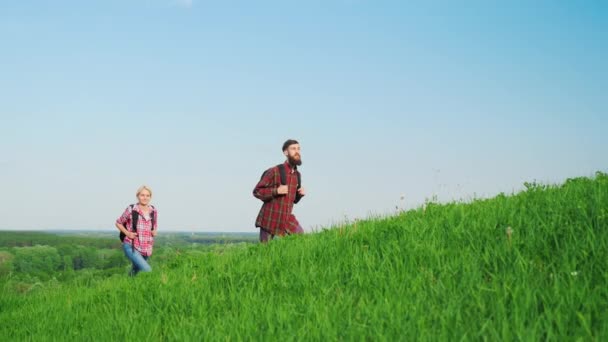 Mężczyzna i kobieta z plecakami idą się stok, zielone wzgórza. W malowniczym miejscu. Aktywny sposób życia, zielonej turystyki. Film w zwolnionym tempie — Wideo stockowe