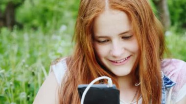 Doğada bir smartphone zevk Kızıl saçlı genç kız gülümsüyor. Parkta yeşil ağaçlar bir arka plan üzerinde oturma portre